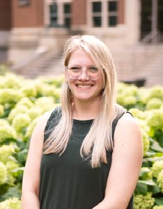 Ariel Steele poses by hydrangeas. Ariel has long blonde hair and wears glasses. She is wearing a dark green sleeveless top