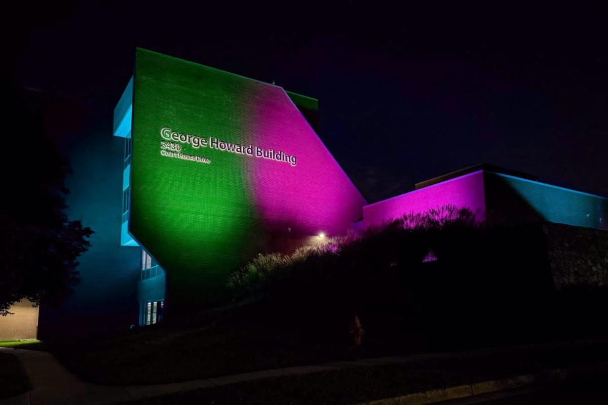Outside of the George Howard Building at night with the side of the building cast in a teal, green, and pink glow.