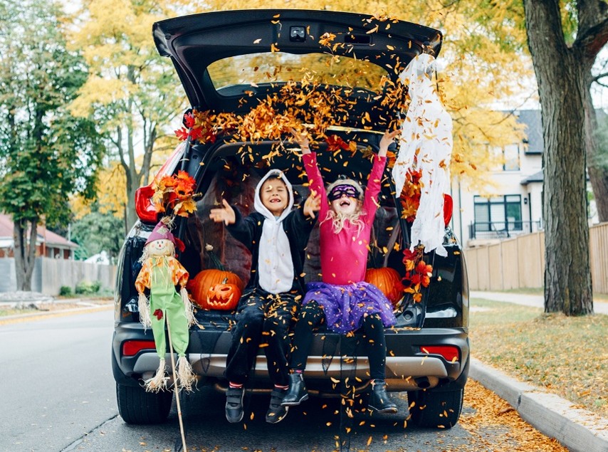 A car parked in a street with its trunk popped open with two children seated inside wearing Halloween costumes and throwing leaves into the air. The trunk is decorated with Halloween decor including a ghost, jack-o'-lantern, and a scarecrow.