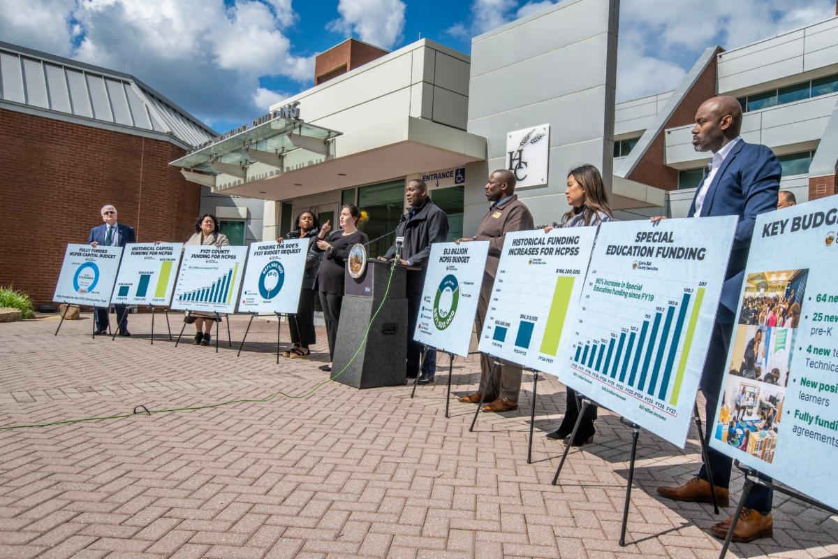 County Executive Ball standing at a podium outside the George Howard Building announcing funding he's included in his proposed Fiscal Year 2027 Operating Budget for Howard County Public School System. Ball is flanked by boards highlighting the funding.