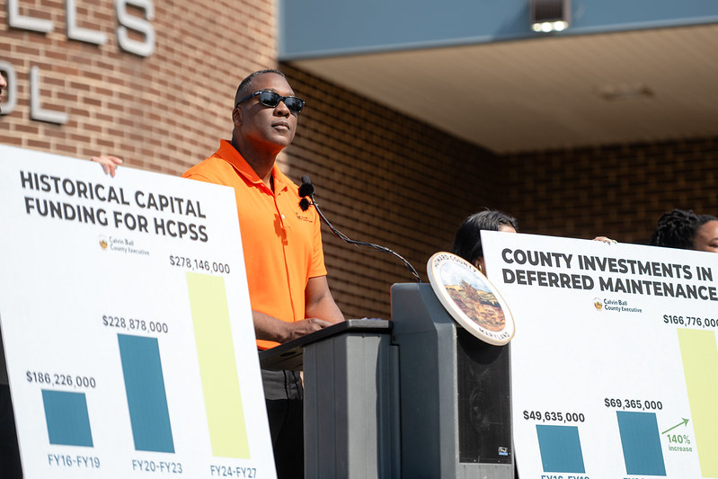 County Executive Calvin Ball speaking at a podium out front of Oakland Mills Middle School at his press conference announcing he will fully fund the Howard County Public School System and its Board of Education's Fiscal Year 2027 Capital Budget requests.