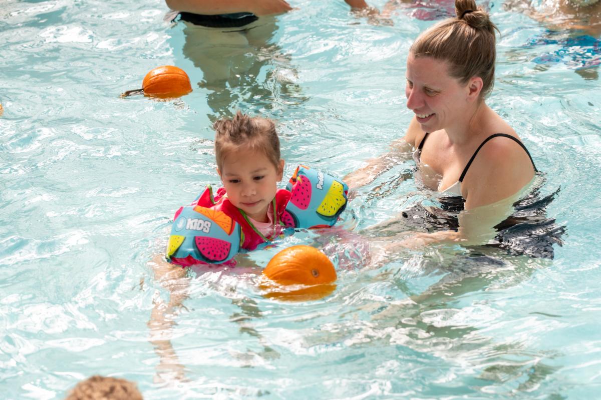 Adult and child floating in a pool with floating pumpkins surrounding in the water.