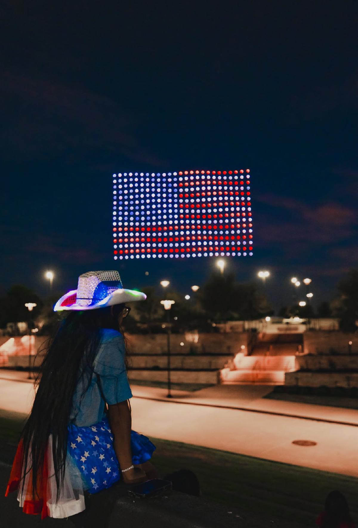 Person dressed up in red, white, and blue in a cowboy hat looking out towards an lite up American Flag made up of hundreds of drones flying together in the night sky.