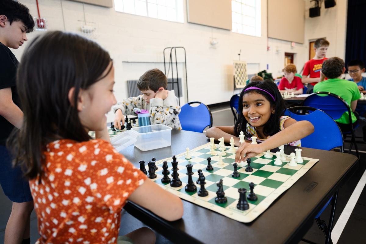Children seated at tables playing chess against one another while other children stand nearby watching.