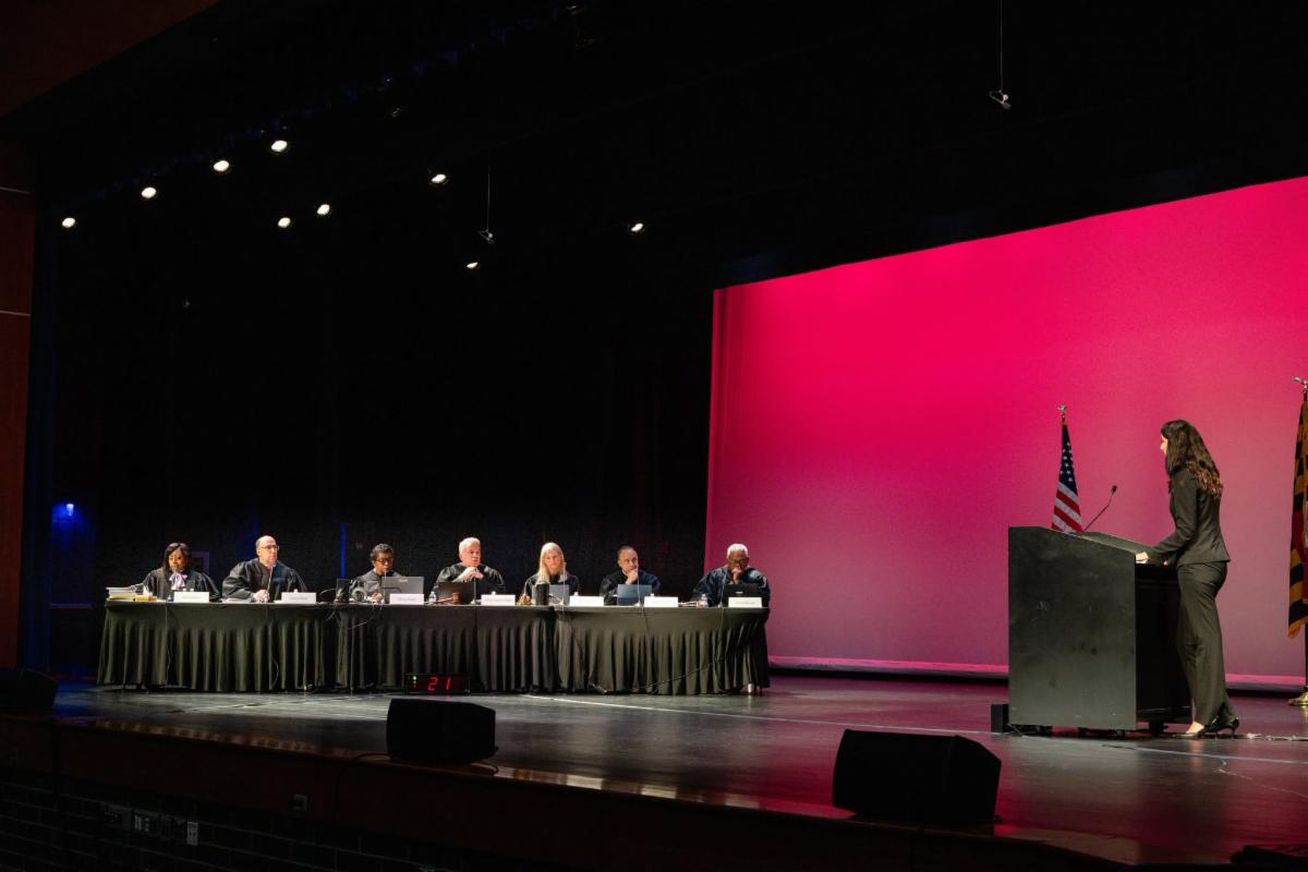 A person standing at a podium on stage at Guilford Park High School facing a table where members of Maryland Supreme Court are seated.