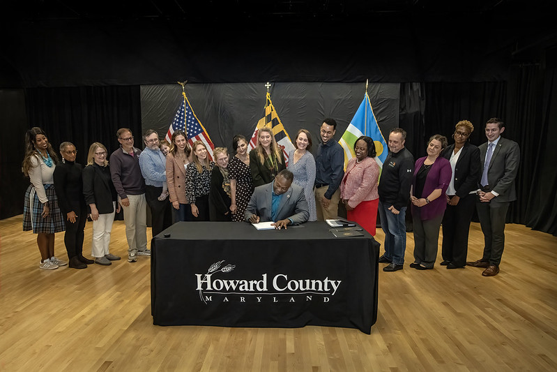 County Executive Calvin Ball seated at a table signing Council Resolution 159-2026 into law as individuals lined up behind him look on. The American, Maryland, and Howard County flags stand in the background.