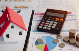 Paperwork laid out on a table with pie charts, bar graphs, line graphs, and tables. On top of the paperwork is a tiny toy house, a calculator, and coins. Some coins are stacked.