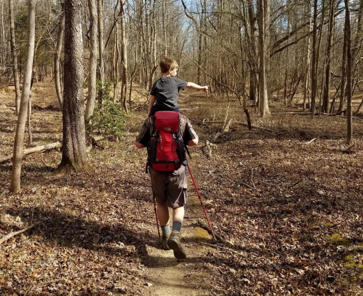 Adult walking through the woods along a trail carrying a child on their shoulders.