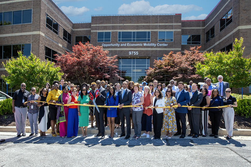County Executive Calvin Ball posed out front of the new Opportunity and Economic Mobility Center at 9755 Patuxent Woods Drive in Columbia holding scissors and surrounded by event attendees as he gets ready to cut the yellow ribbon opening the building.