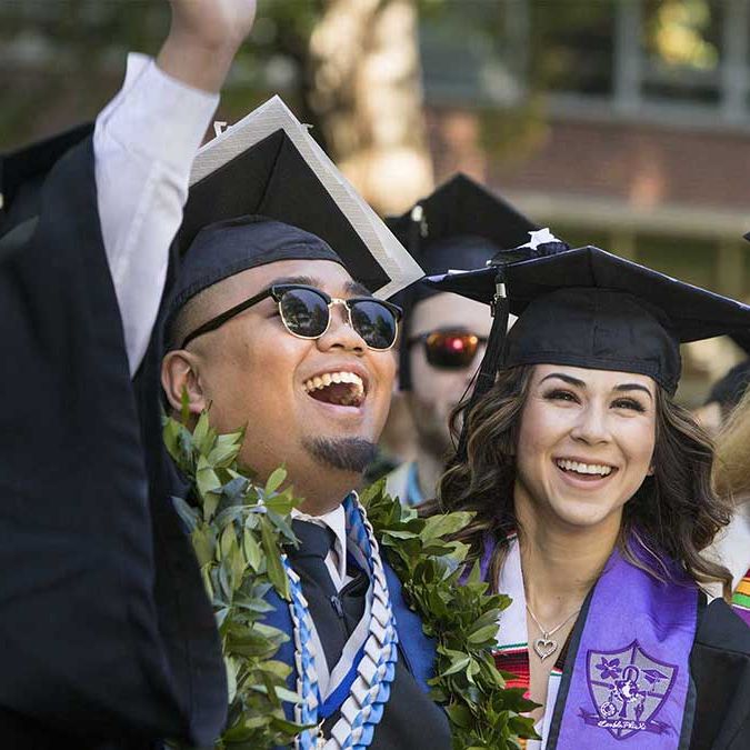 Graduates smile at commencement in caps, gowns and flower leas. One pumps his fist in the air in triumph.