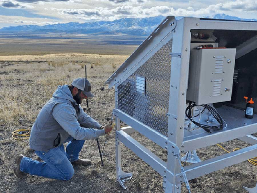 Person adjusting equipment at a weather monitoring station in an open field with mountainous background.