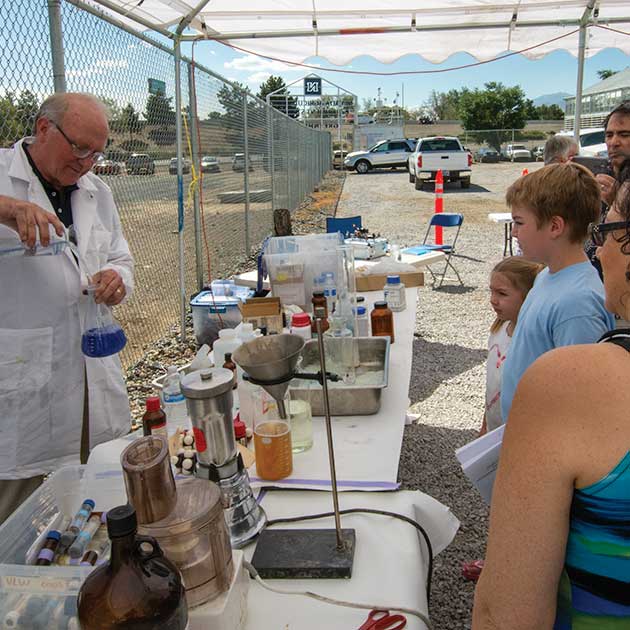 Glenn Miller wearing a stained lab coat and pouring a bright blue liquid into a beaker as kids and parents look on with curiosity.