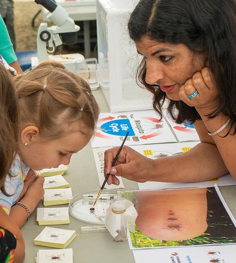 Monika Gulia-Nuss at Field Day showing her tick research, with insects, petri dishes, microscopes, photos and more, to two young girls.