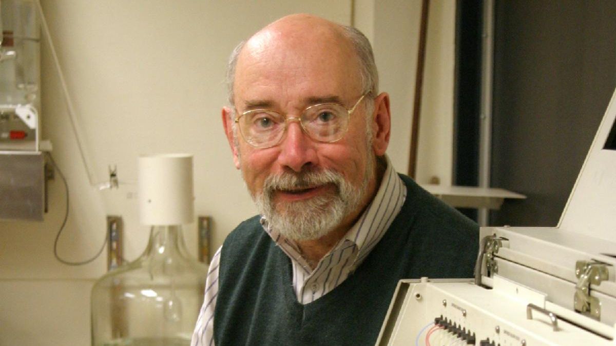 A man in glasses sitting by a wired lab equipment.