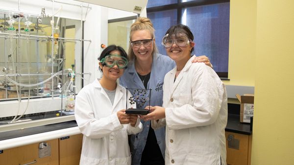 Lyndsay Munro smiles with her arms around two students who are holding a model. They are wearing PPE and are standing in front of a laboratory fume hood.