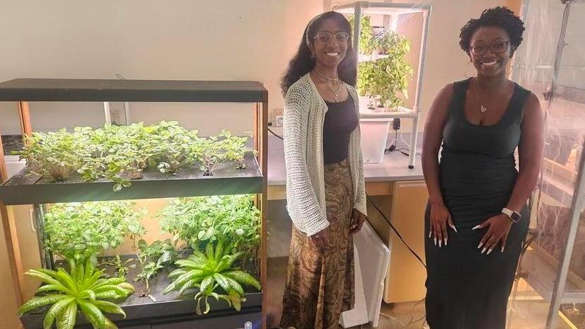 Two smiling women stand beside a glass container filled with growing plants.