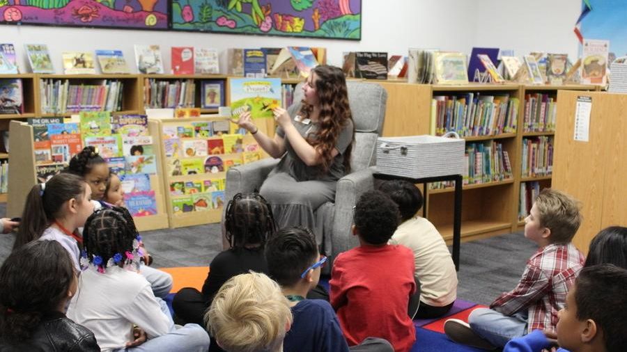 A woman speaks to a group of children seated on the floor in front of shelves filled with colorful books.