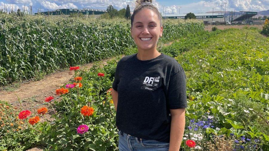 A smiling woman poses amid a farm with green plants and flowers.