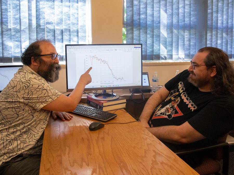 David Alvarez-Ponce and James Vesper sit on opposite sides of a desk, while Alvarez-Ponce points to his computer screen.