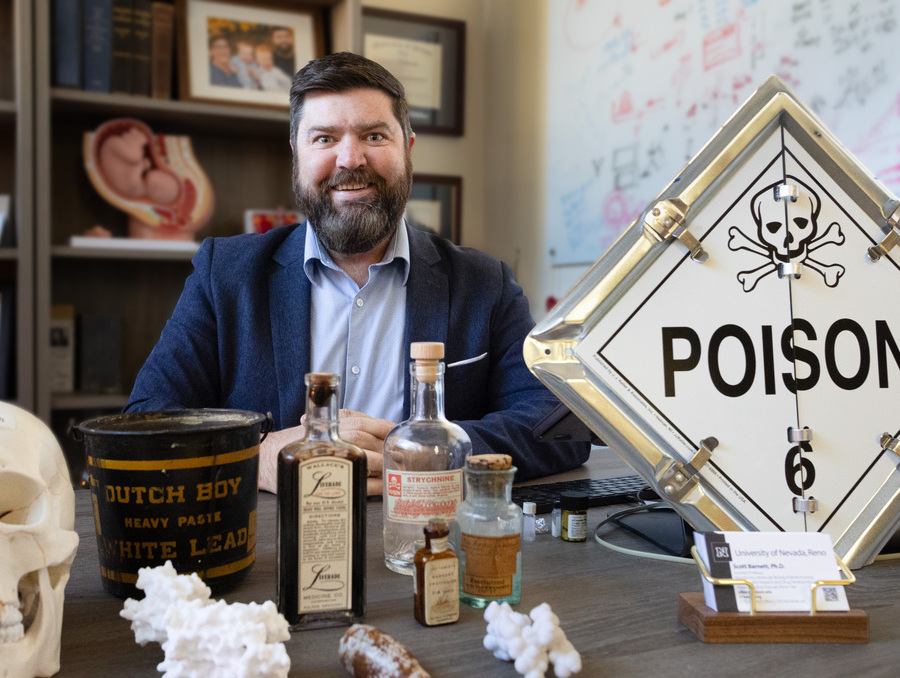 Scott Barnett sitting at a desk with antique poisonous liquids preserved in labeled bottles. 