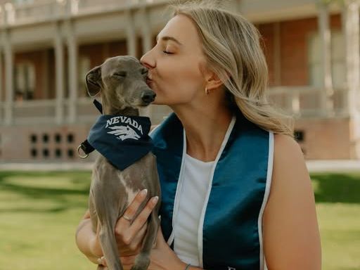A person in a white outfit and graduation stole is holding a small dog wearing a "Nevada" bandana, standing outside a building on a grassy area. The person is gently kissing the dog's head.