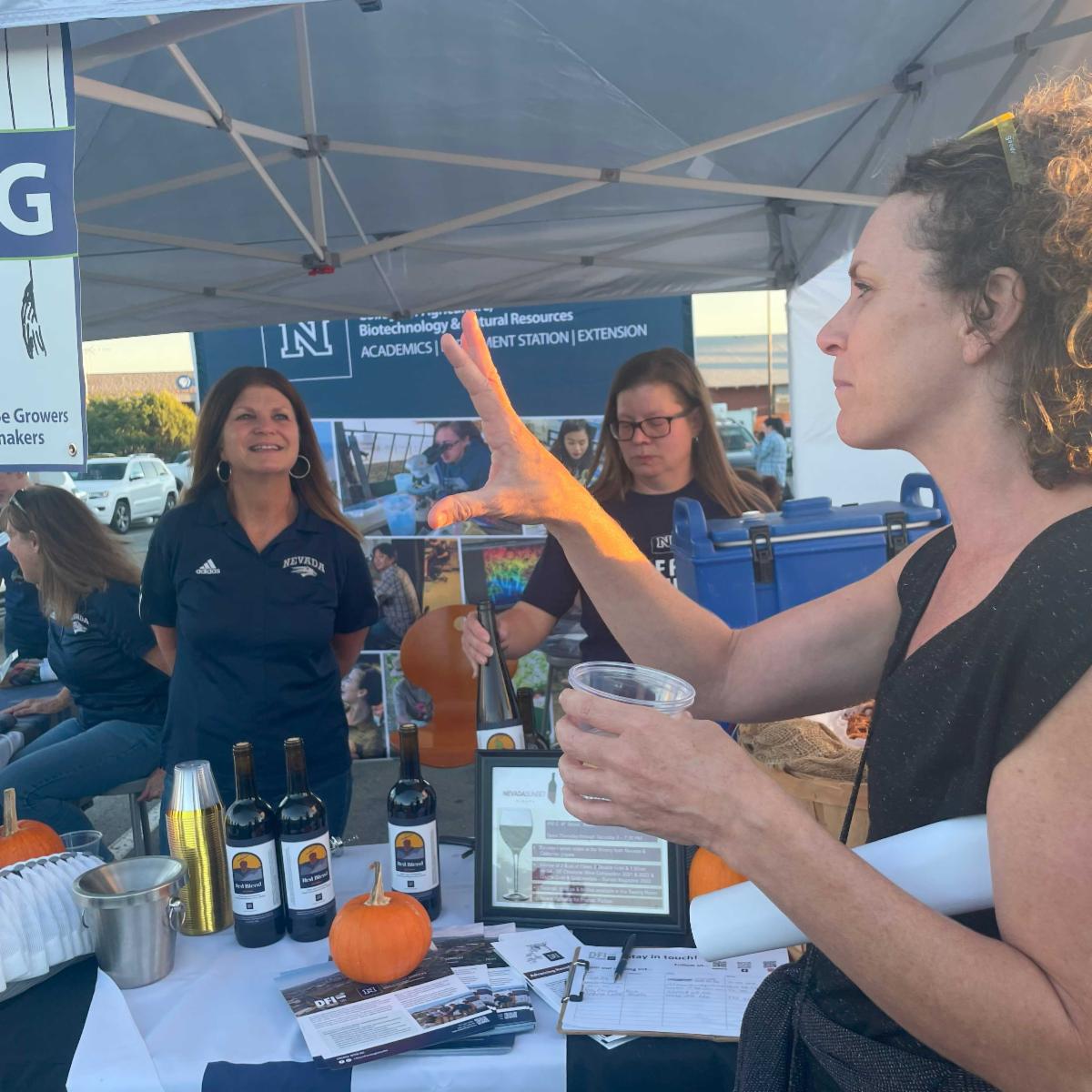 Claudene Wharton staffing a wine tasting booth at which a community member is sampling the University's white wine.