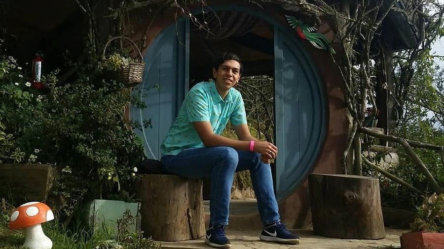 A man sits on a tree log in front of a round blue door that leads into a house with a culturally styled exterior.