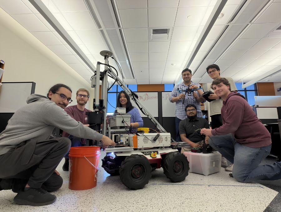 Parikshit and students pose around a robot on wheels inside a classroom. 