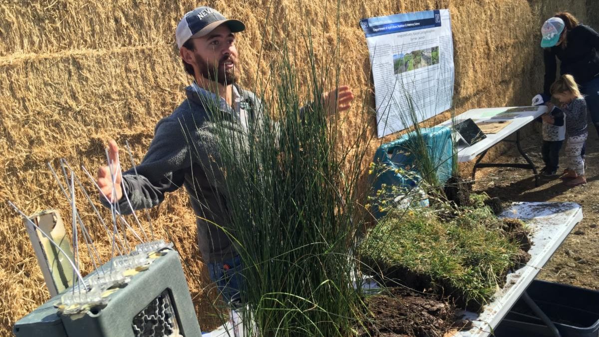 A researcher at a table of grasses and research equipment holds his hands up as if to say "The fish was this big."