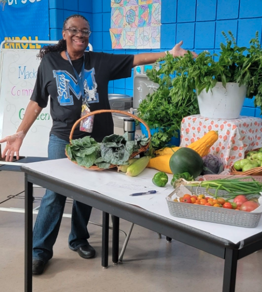 Educator Shirley Brezzell stands with her arms stretched wide by a table of produce.