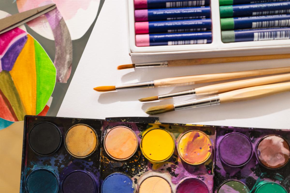 Close-up image of watercolor paint pans with vivid splashes, paintbrushes, and pastel sets in the background