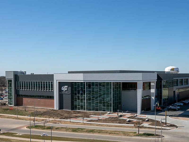The front of the NIAR Hub for Advanced Manufacturing and Research under bright blue skies with construction material around the perimeter of the building. A water tower with an illustration of WuShock on is in the background poking above the building