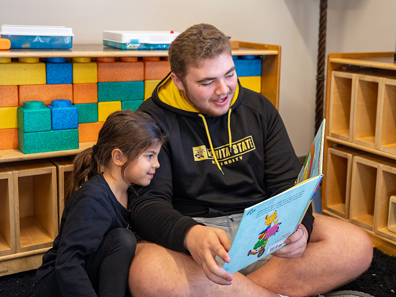 A WSU student teacher in a Wichita State University hoodie sits on the floor to read a children's book to a kindergartner in a class. The background has multiple shelves with various building blocks and school supplies.