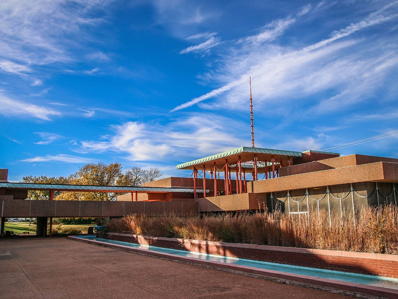 The Corbin Education Center is pictured undy partly cloudy blue skies. The water fountain is in the foreground with the rest of the building and one of the spires in the background