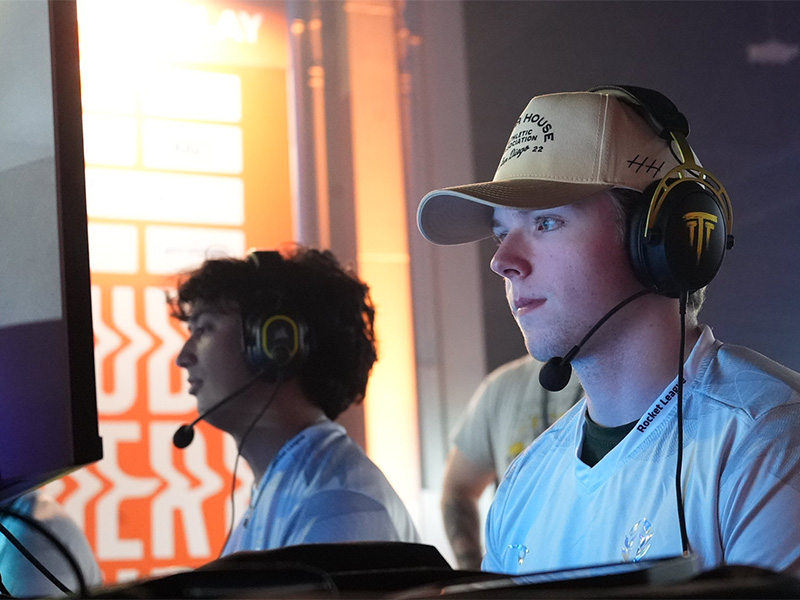 Two students in Wichita State's esports program sit at a computer desk, both with headsets on, to compete with another university in a Rocket League tournament