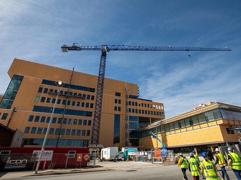 A view of the under construction Wichita Biomedical Campus from the ground. Photo shows the outer walls made out of orange construction material. On the ground is various equipment and vehicles for the construction, and workers in hardhats and high visibility vests are also shown. Centered in the photo is a large blue crane used during the construction process.