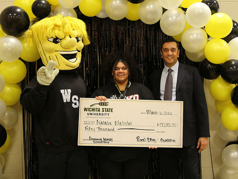 Natalie Malcolm poses with her $50,000 scholarship check in front of an arch of yellow, white and black balloons and a black curtain, to her right is WuShock giving the WSU "Shocker" hand sign and to her left is Ronal Bhagat in a black suit and gray checkerboard tie