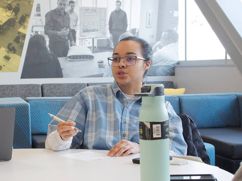 Wichita State junior Maya Douglas, in a blue and white button up, talks to someone off screen as she writes something on a piece of paper. A water bottle and smartphones sit on the table in front of her.