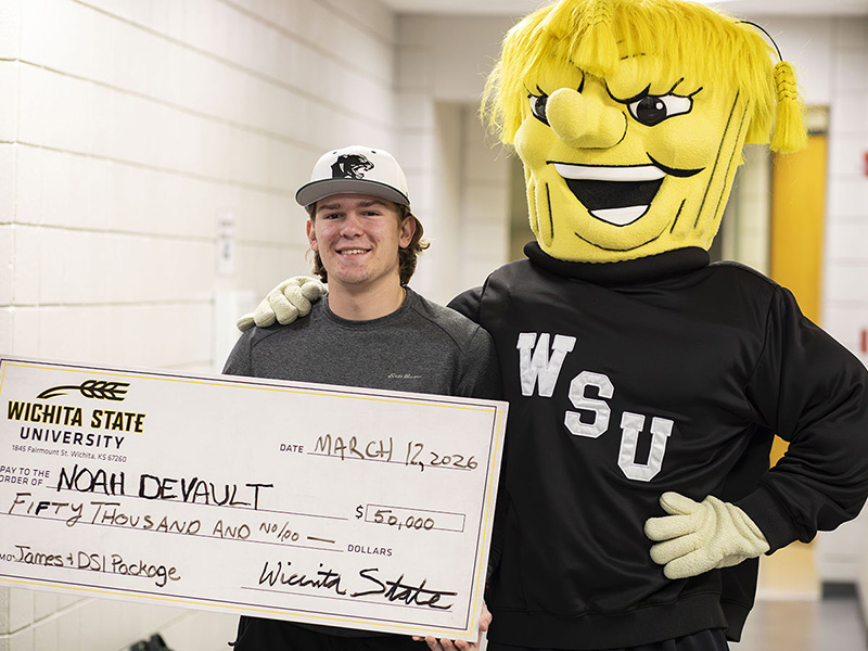 Noah DeVault poses with his $50,000 scholarship check alongside WuShock in a hallway with white brick walls at Derby High School