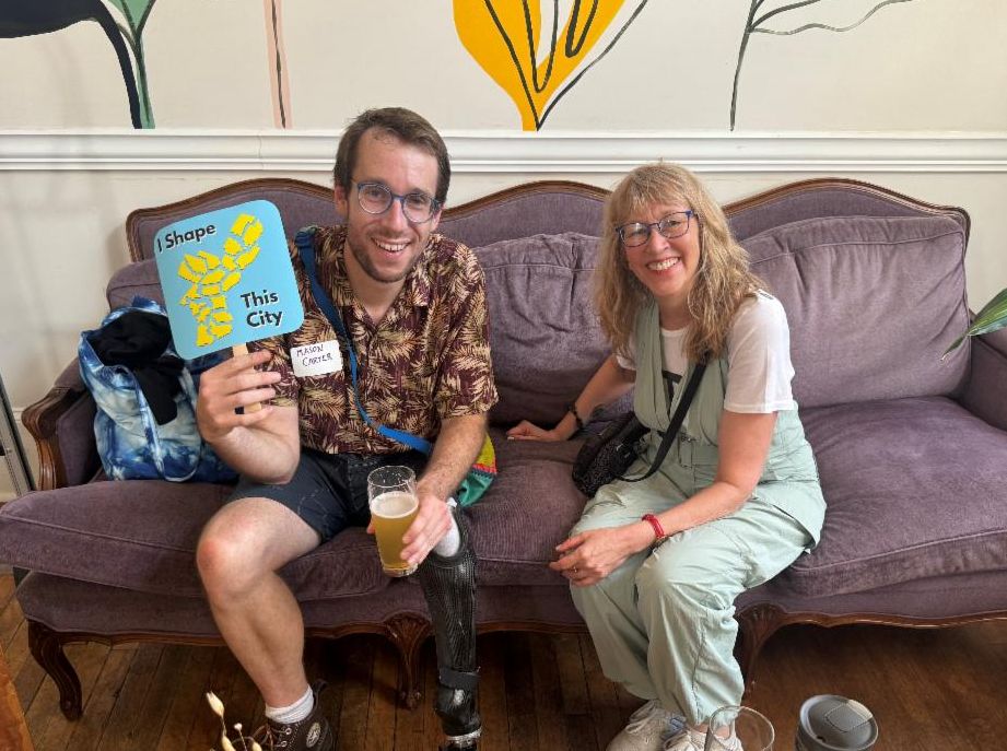 Photo of Donna and Mason sitting on a purple couch. Mason is holding an "I shape this city" blue fan.