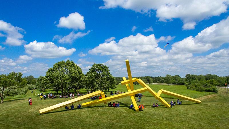 A large yellow sculpture of long lines and tubes in no particular shape sits on a grassy lawn in the sun. It is much larger than the people looking at it.