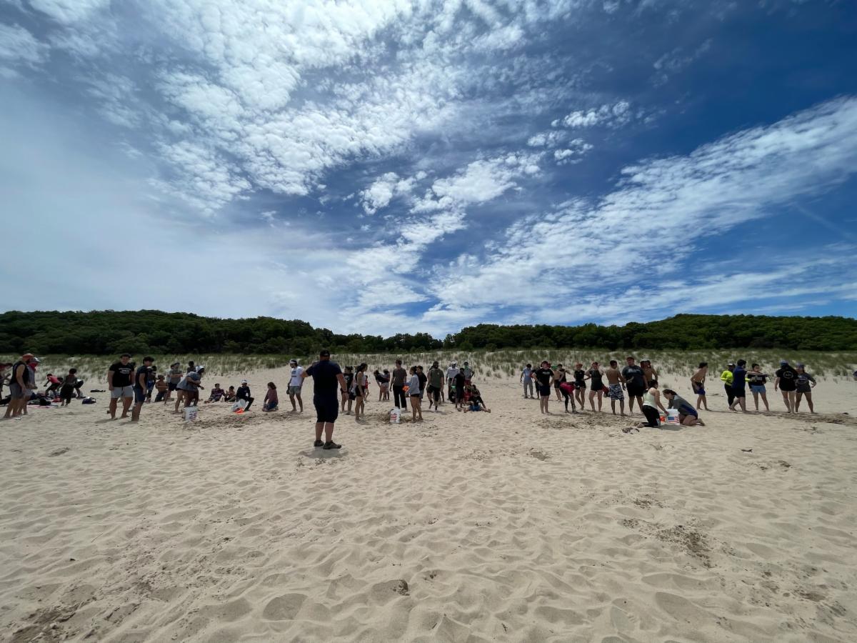 A large group of teenagers revels in the sand on a sunny beach under bright blue skies