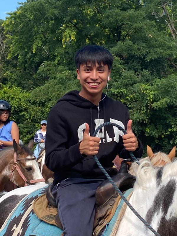 A young man sits on a horse and flashes a big smile and thumbs up to the camera.