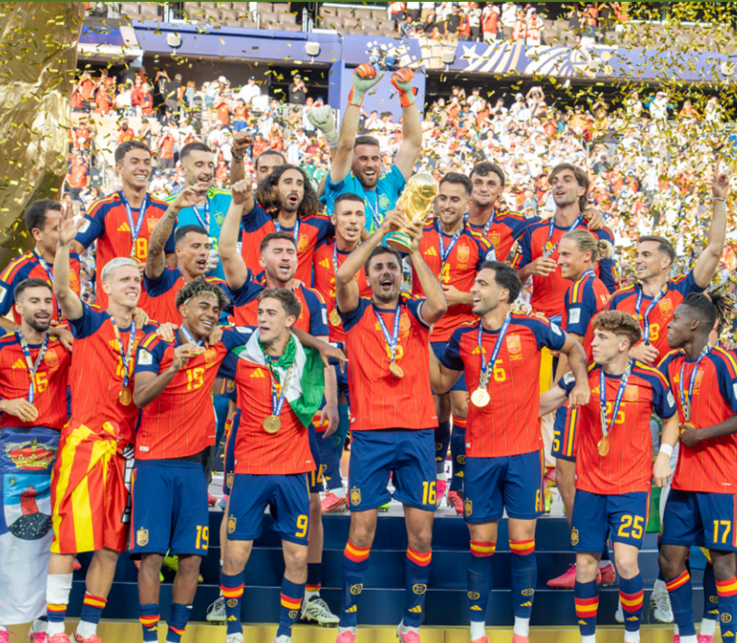 A group of soccer team players wearing red, yellow, and blue cheer in a soccer stadium holding the World Cup trophy as fans cheer and confetti falls in the background