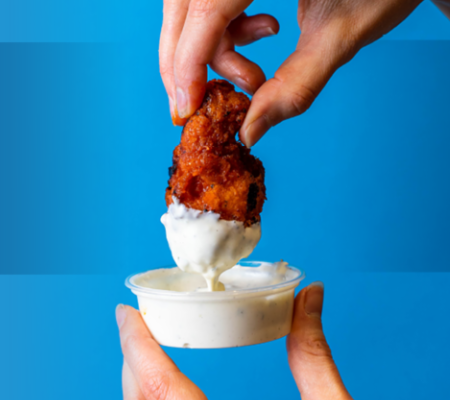 A pair of hands hold a ranch cup, and dips a chicken wing into the ranch in front of a blue background