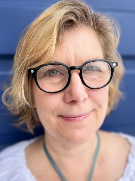 Lauren Wyeth headshot. Lauren is a white woman with short blond hair and blue eyes. She is wearing dark framed glasses a blue necklace and a white top and is pictured against a blue background.