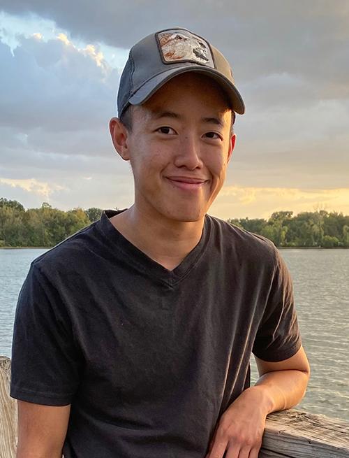 Photo of Jie, a nonbinary Asian person with short black hair. Jie is wearing a short sleeve shirt and baseball cap and standing near a lake.