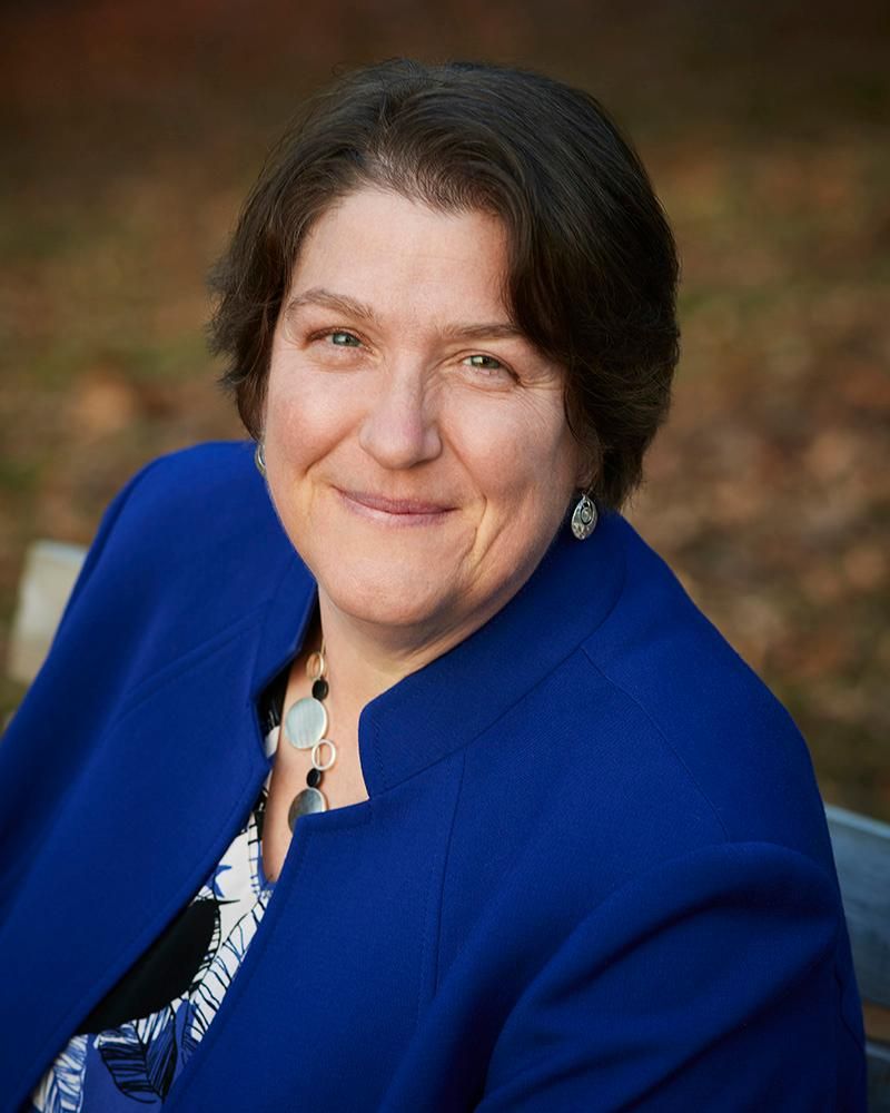 A headshot of Rev. Jen looking at the camera. Rev. Jen is a white woman with short brown hair. She is wearing a blue jacket and earrings and a necklace and is smiling with her mouth closed.