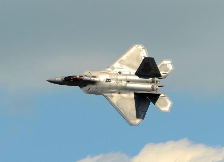 A silver jet plane angles across a blue sky with gray clouds. It is a Lockheed Marting F-22 Raptor military plane.