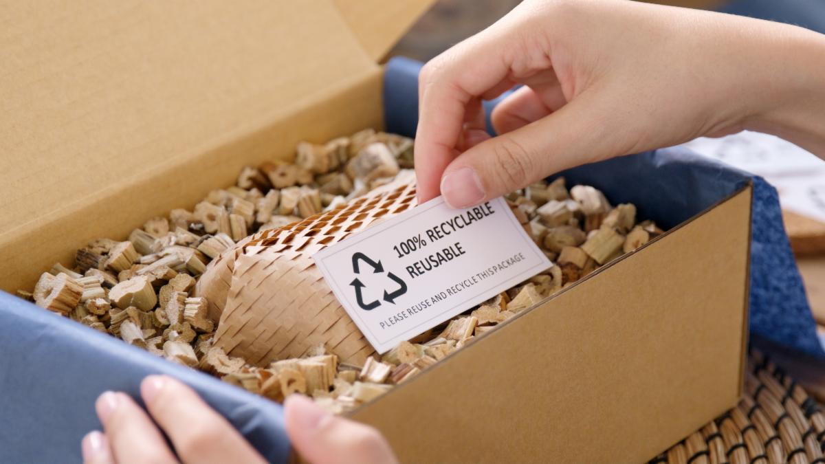 A corrugated cardboard box is filled with brown packaging filler. A pair of hands holds the box.